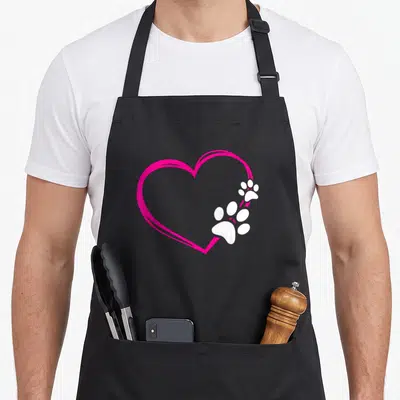 Tablier de cuisine noir avec un grand cœur rose vif et deux empreintes de pattes blanches superposées sur le côté droit du dessin.