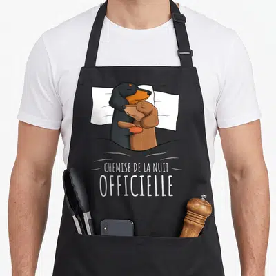 Tablier de cuisine noir avec deux teckels s'enlaçant dans un lit et le texte « chemise de la nuit » imprimé en blanc.