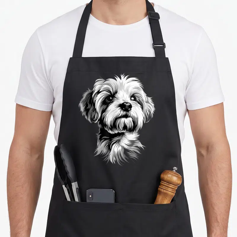 Tablier de cuisine noir avec illustration détaillée d'un Bichon Maltais blanc au regard expressif et aux poils volumineux.