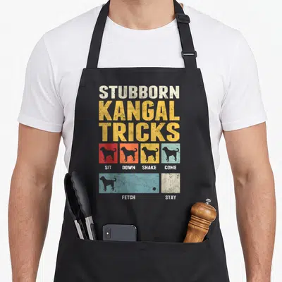 Tablier de cuisine noir avec inscription Stubborn Kangal Tricks et silhouettes de chiens colorées.