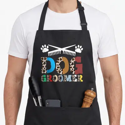Tablier de cuisine noir avec inscription Dog Groomer colorée, peigne blanc et traces de pattes sur fond uni.