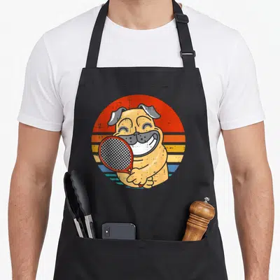 Tablier de cuisine noir avec illustration d'un bouledogue souriant tenant une raquette de tennis rouge sur fond circulaire orange et bleu.