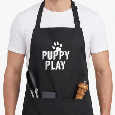 Tablier de cuisine noir avec l'inscription Puppy Play et une patte de chien blanche au centre du torse.
