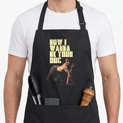 Tablier de cuisine noir avec texte Now I Wanna Be Your Dog et illustration d'Iggy Pop en position quadrupède aux teintes orangées.