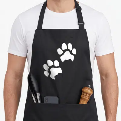 Tablier de cuisine noir avec deux grandes empreintes de pattes de chien blanches imprimées au centre du vêtement.
