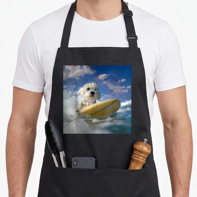 Tablier de cuisine noir avec un Coton de Tuléar blanc sur une planche de surf jaune dans l'eau salée.