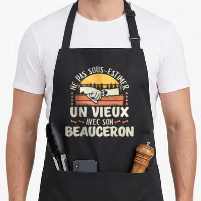 Tablier de cuisine noir avec illustration vintage d'une poignée de main entre main humaine et patte de chien Beauceron sur fond de soleil orange.