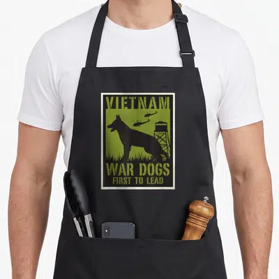 Tablier de cuisine noir avec silhouette de chien, hélicoptères et texte Vietnam War Dogs First to Lead sur fond vert olive.