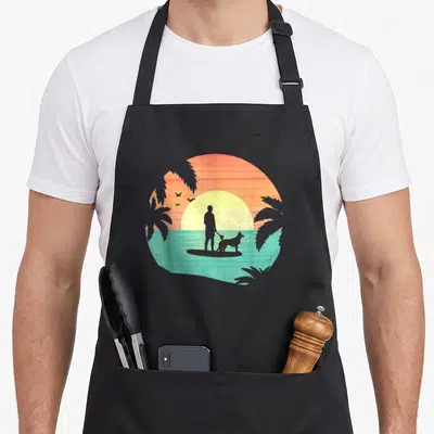 Tablier de cuisine noir avec illustration d'un homme et d'un chien en stand up paddle au coucher du soleil tropical.
