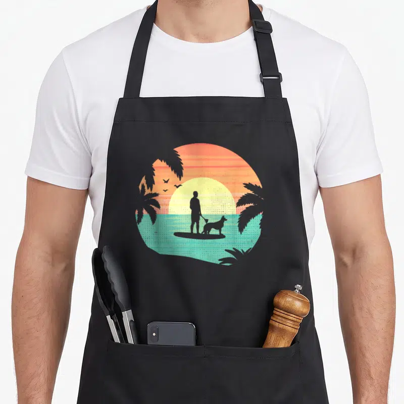 Tablier de cuisine noir avec illustration d'un homme et d'un chien en stand up paddle au coucher du soleil tropical.