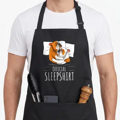 Tablier de cuisine noir avec deux bulldogs anglais s'enlaçant sur un lit et le texte Official Sleepshirt en blanc.