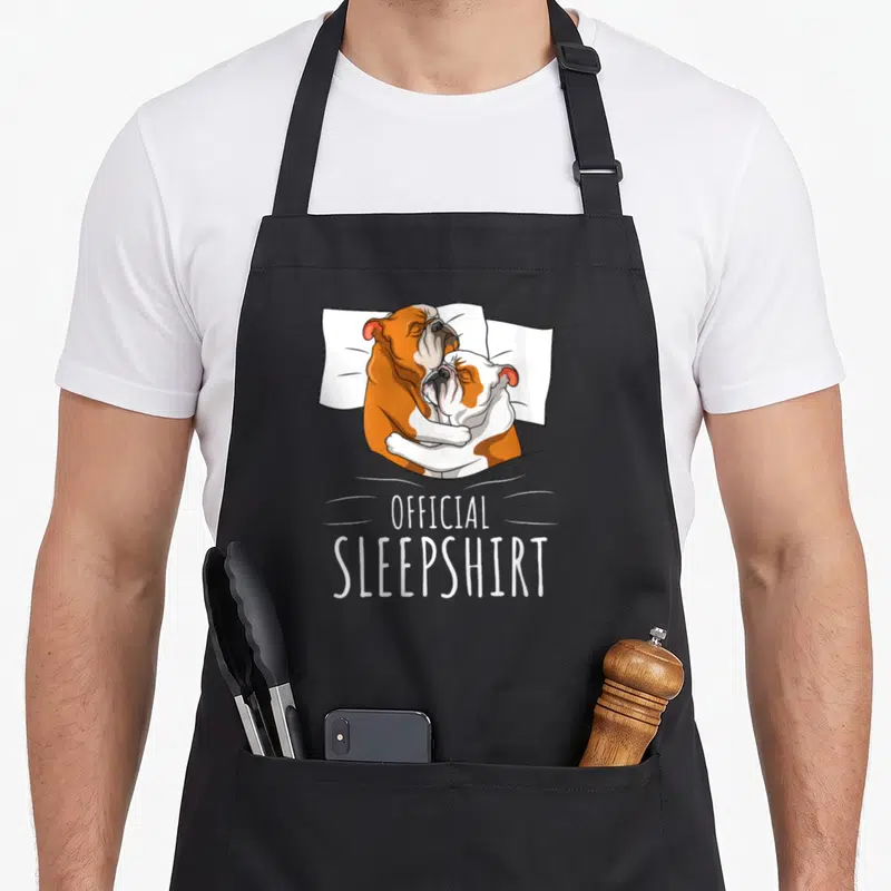 Tablier de cuisine noir avec deux bulldogs anglais s'enlaçant sur un lit et le texte Official Sleepshirt en blanc.