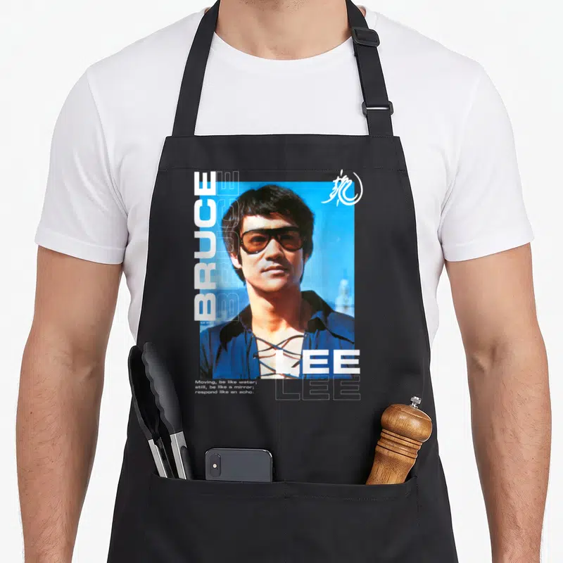Tablier de cuisine noir avec portrait de Bruce Lee, lunettes et texte Moving be like water en typographie blanche.