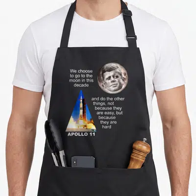 Tablier de cuisine noir avec illustration du lanceur Apollo 11 et portrait circulaire de John F. Kennedy sur fond gris.