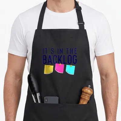 Tablier de cuisine noir avec le texte It's in the backlog et trois post-its jaune, rose et bleu.