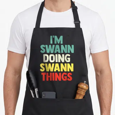Tablier de cuisine noir avec inscription I'M SWANN DOING SWANN THINGS en typographie multicolore.