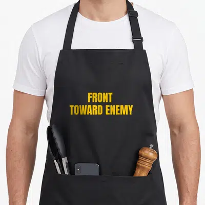Tablier de cuisine noir avec l'inscription Front Toward Enemy en lettres jaunes capitales centrées sur le devant.