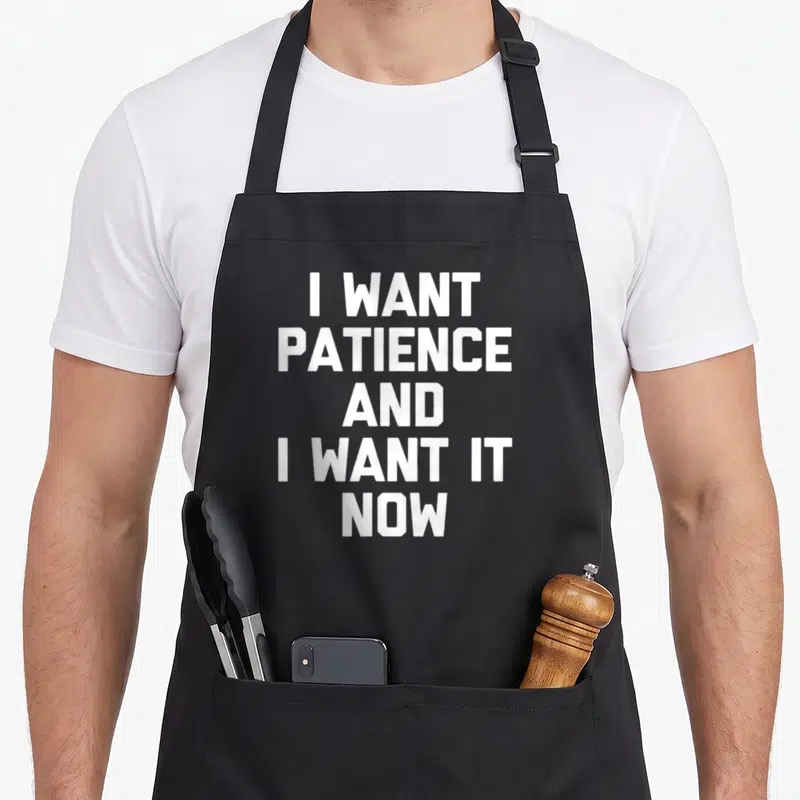 Tablier de cuisine noir avec le texte blanc en majuscules I Want Patience And I Want It Now centré sur le devant.