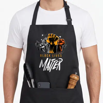 Tablier de cuisine noir avec plusieurs poings levés colorés et le texte Black Lives Matter imprimé en orange et blanc.
