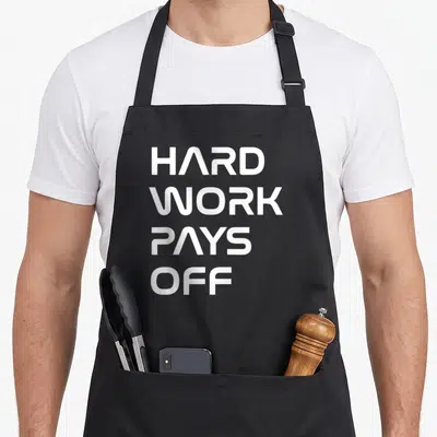 Tablier de cuisine noir avec l'inscription Hard Work Pays Off en lettres blanches capitales stylisées sur fond uni.