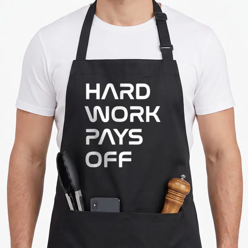 Tablier de cuisine noir avec l'inscription Hard Work Pays Off en lettres blanches capitales stylisées sur fond uni.