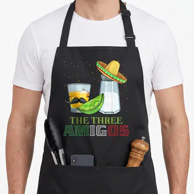 Tablier de cuisine noir avec illustration de verre de tequila à moustache, lime, perruche et texte The Three Amigos.