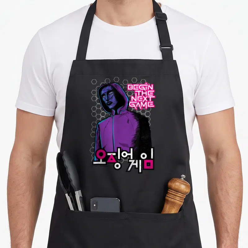Tablier de cuisine noir avec un personnage hoodé violet et le texte Begin The Next Game en rose.
