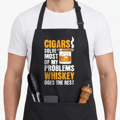 Tablier de cuisine noir avec un verre de whiskey et un cigare fumant au-dessus du texte imprimé.