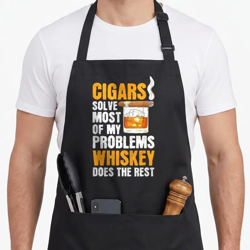 Tablier de cuisine noir avec un verre de whiskey et un cigare fumant au-dessus du texte imprimé.