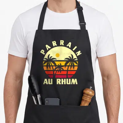Tablier de cuisine noir avec illustration soleil couchant palmiers et texte parrain au rhum en style rétro.