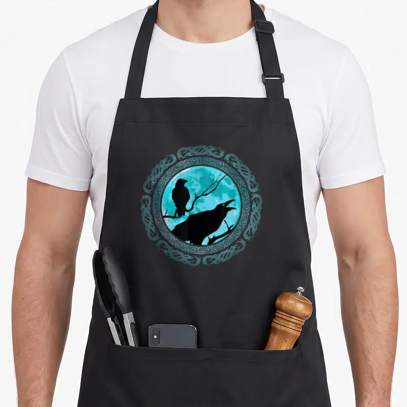 Tablier de cuisine noir avec illustration circulaire de deux corbeaux noirs sur fond bleu turquoise et cadre de nœuds celtiques.