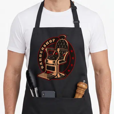 Tablier de cuisine noir avec illustration d'une chaise de barbier vintage et l'inscription BARBERSHOP sur fond circulaire rouge et noir.