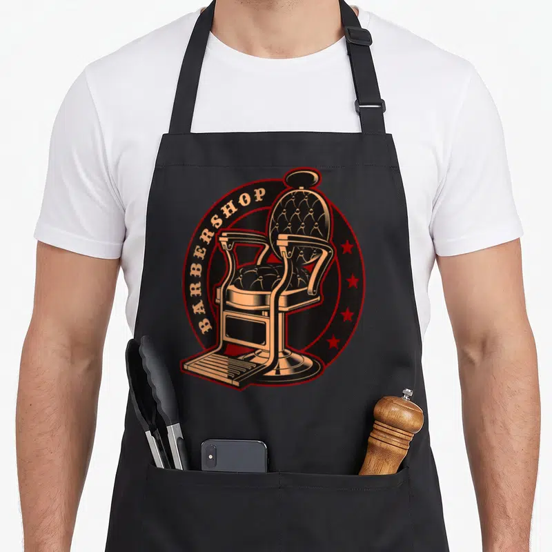 Tablier de cuisine noir avec illustration d'une chaise de barbier vintage et l'inscription BARBERSHOP sur fond circulaire rouge et noir.