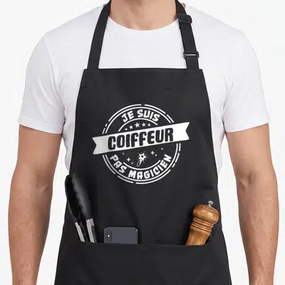 Tablier de cuisine noir avec un logo circulaire blanc affichant Je suis coiffeur pas magicien et une étoile filante.