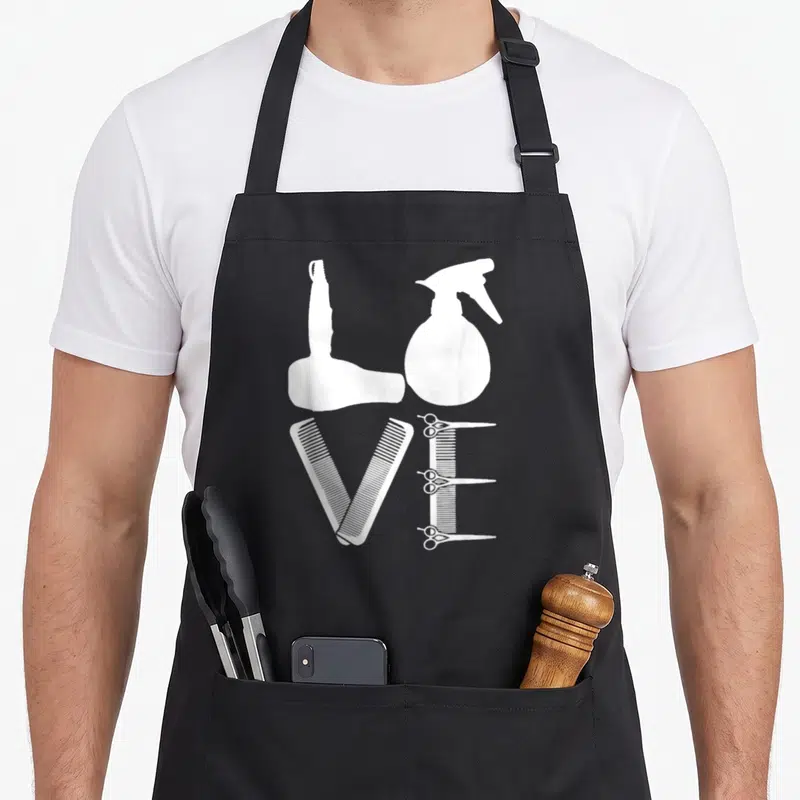 Tablier de cuisine noir avec les mots LOVE formés par un séchoir, une bouteille pulvérisateur, des peignes et des ciseaux blancs.