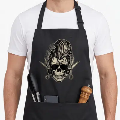 Tablier de cuisine noir avec crâne rockabilly coiffé pompadour et ciseaux de barbier croisés.