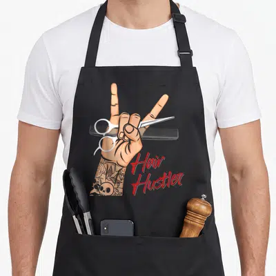 Tablier de cuisine noir avec main tatouée tenant ciseaux et peigne, texte Hair Hustler en rouge.