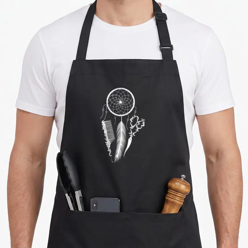 Tablier de cuisine noir avec un capteur de rêves, une peigne et des ciseaux stylisés en blanc.