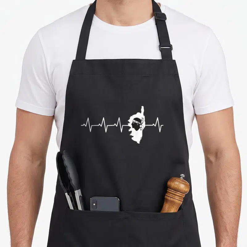 Tablier de cuisine noir avec silhouette corse et ligne de battement de cœur blanche stylisée.