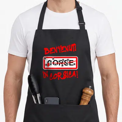 Tablier de cuisine noir avec linscription Benvenuti in Corsica et une plaque blanche traversée par des impacts de balles rouges.