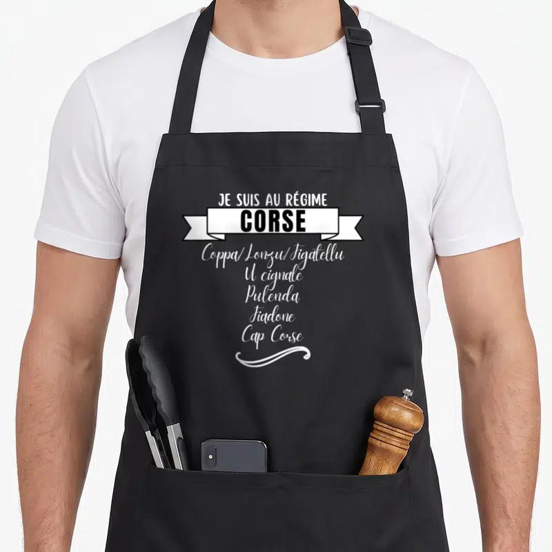 Tablier de cuisine noir avec inscription blanche Je suis au régime Corse et liste de charcuteries traditionnelles sous une bannière décorative.