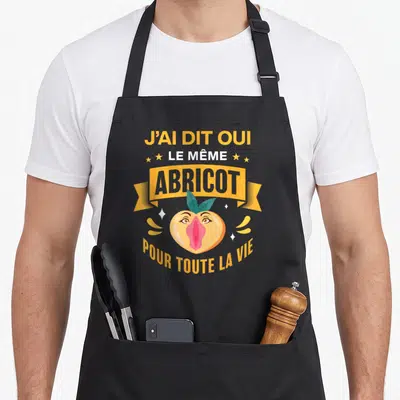Tablier de cuisine noir avec un abricot anthropomorphisé aux yeux rieurs et à la bouche rose, encadré par des banderoles jaunes et blanches.