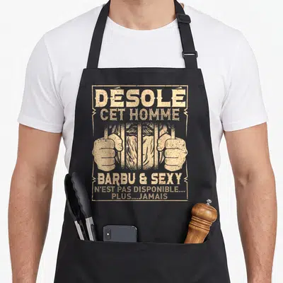 Tablier de cuisine noir avec illustration d'un homme barbu derrière des barreaux et texte beige désolé cet homme barbu et sexy.