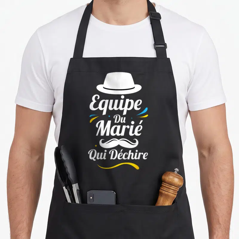 Tablier de cuisine noir avec le texte Equipe Du Marié Qui Déchire, une moustache stylisée et un chapeau blanc.
