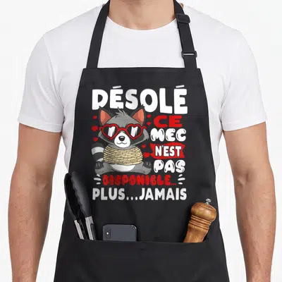 Tablier de cuisine noir avec un raton laveur en lunettes cœur et texte humoristique sur fond uni.