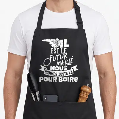 Tablier de cuisine noir avec une main blanche pointant et le texte Il est le futur marié nous sommes juste là pour boire.
