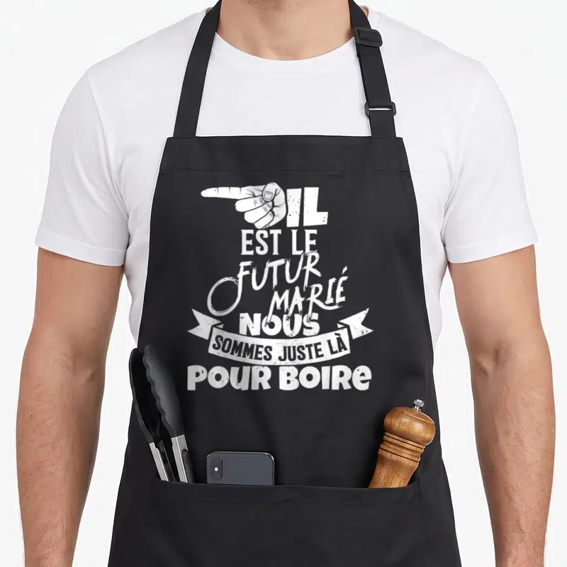 Tablier de cuisine noir avec une main blanche pointant et le texte Il est le futur marié nous sommes juste là pour boire.