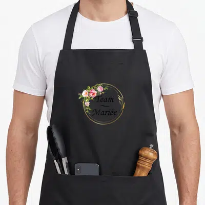 Tablier de cuisine noir avec le texte Team Mariée entouré de fleurs roses et d'un cercle doré élégant.