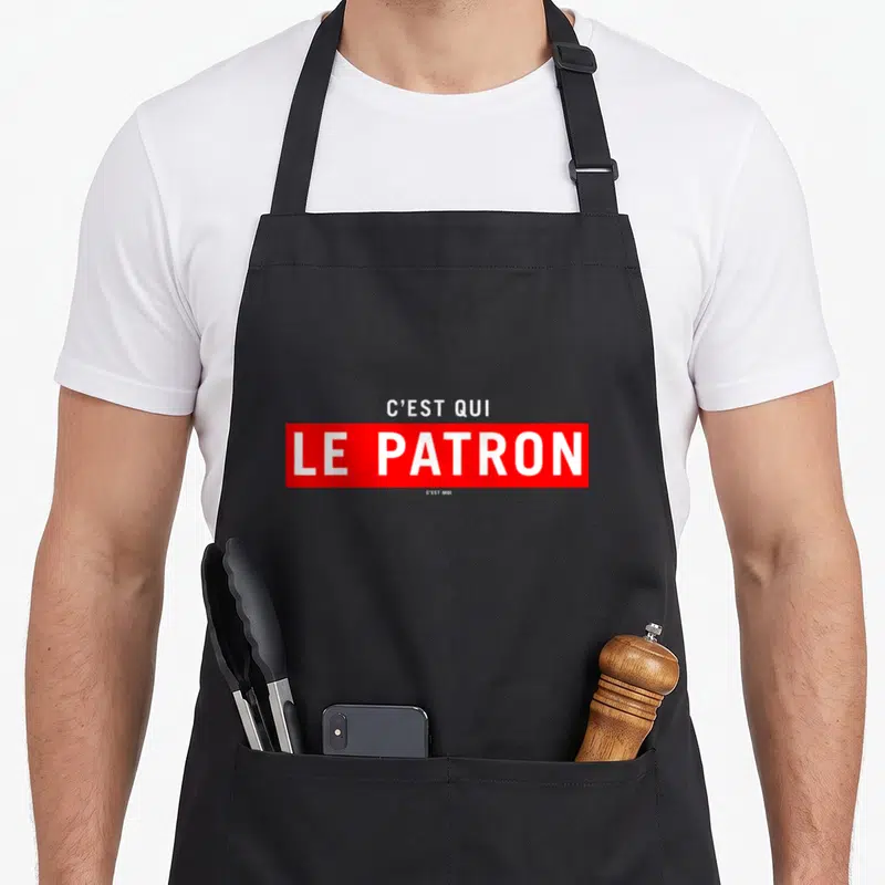 Tablier de cuisine noir avec inscription C'est qui le patron c'est moi en lettres blanches sur fond rouge.
