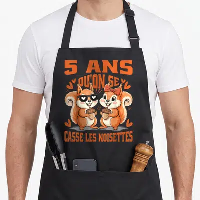 Tablier de cuisine noir avec deux écureuils orange tenant des noix et le texte 5 ans casse les noisettes imprimé en lettres oranges.